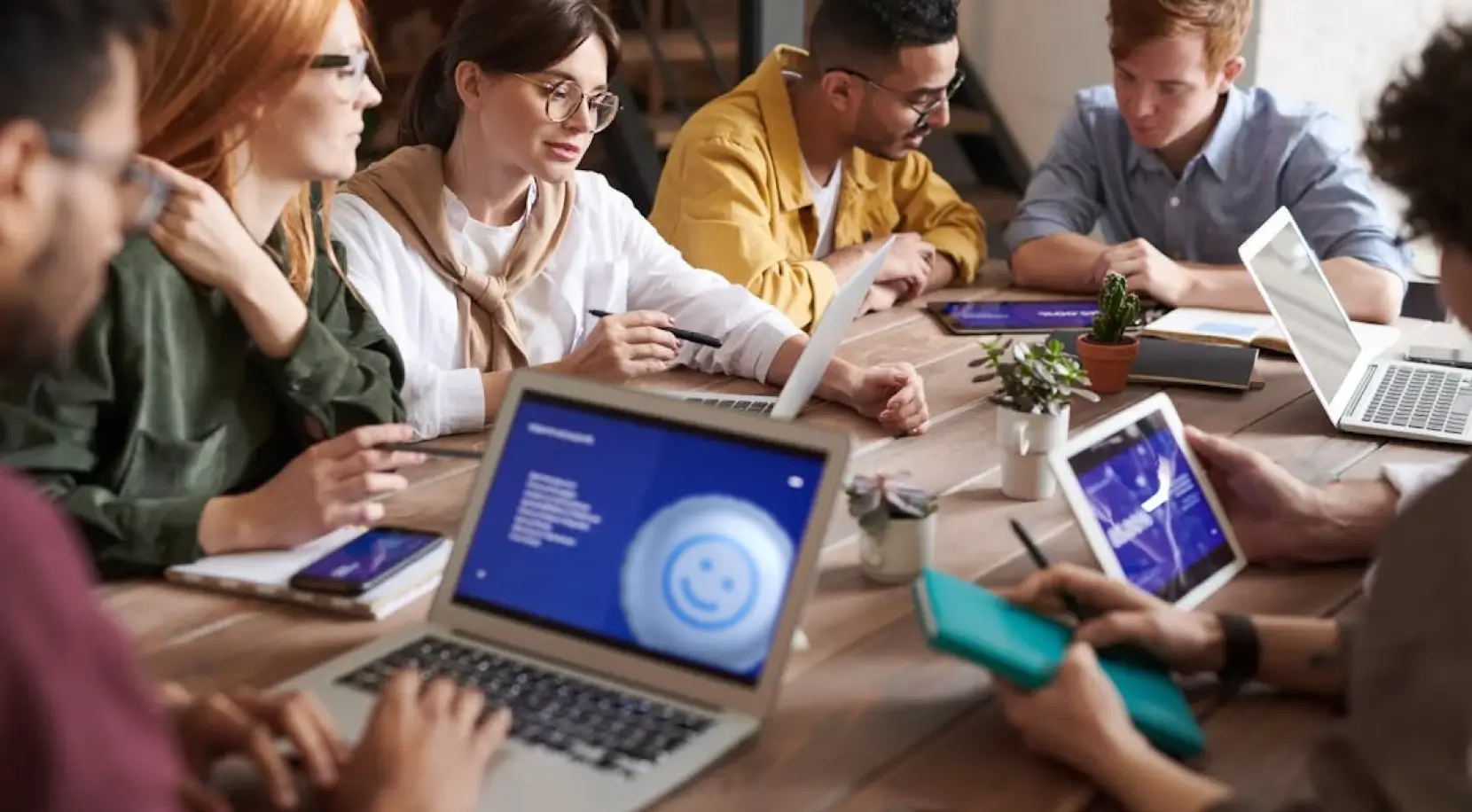 A group of young professionals collaborating around a wooden table with laptops, tablets, and notebooks, engaged in a meeting or discussion in a modern office setting.